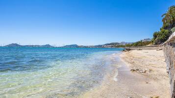 Een strand met helder water, een stenen muur en op de achtergrond bergen.