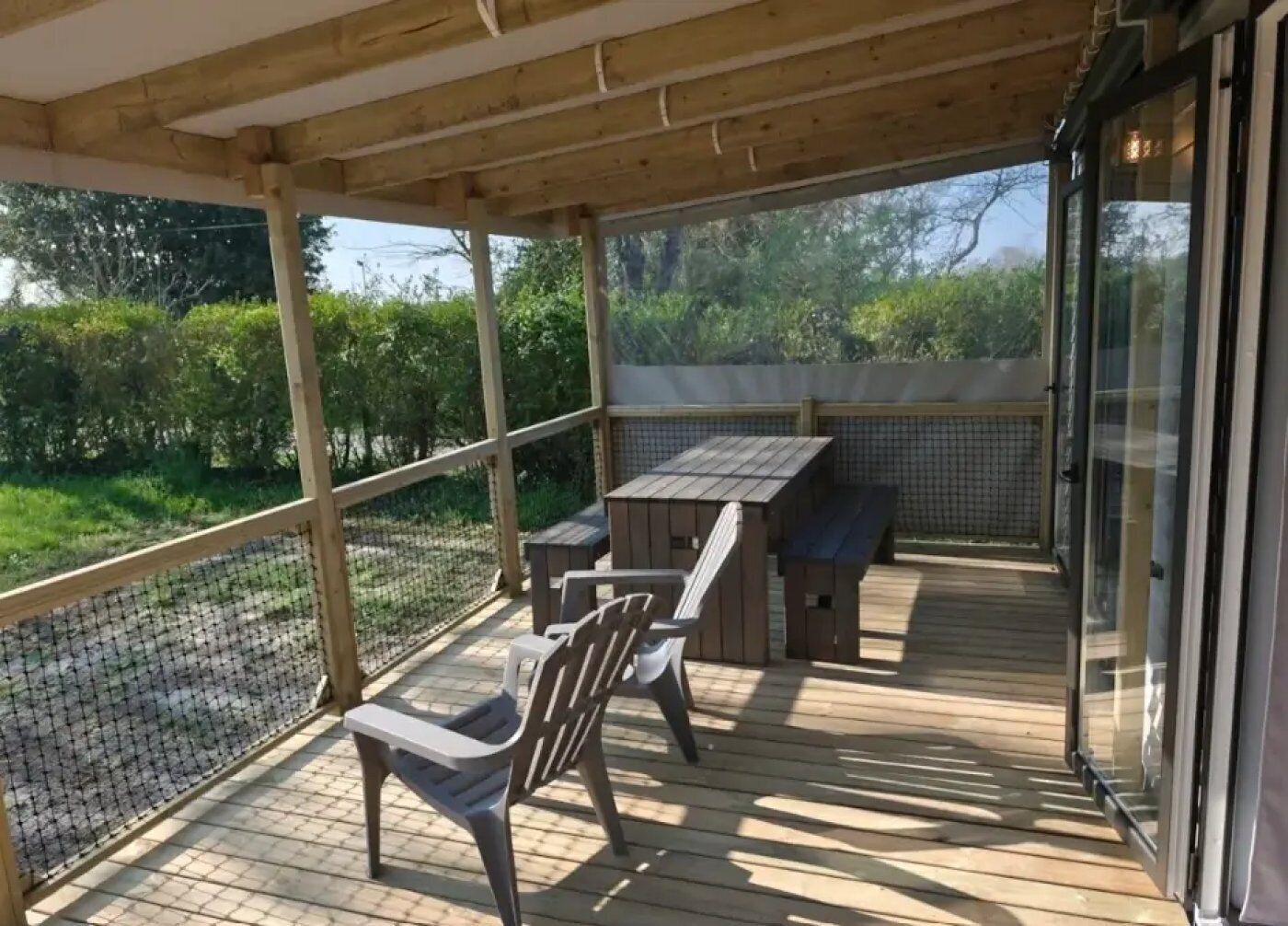 En overdækket terrasse med et bord og stole, omgivet af et træhegn og udsigt til grøn bevoksning.