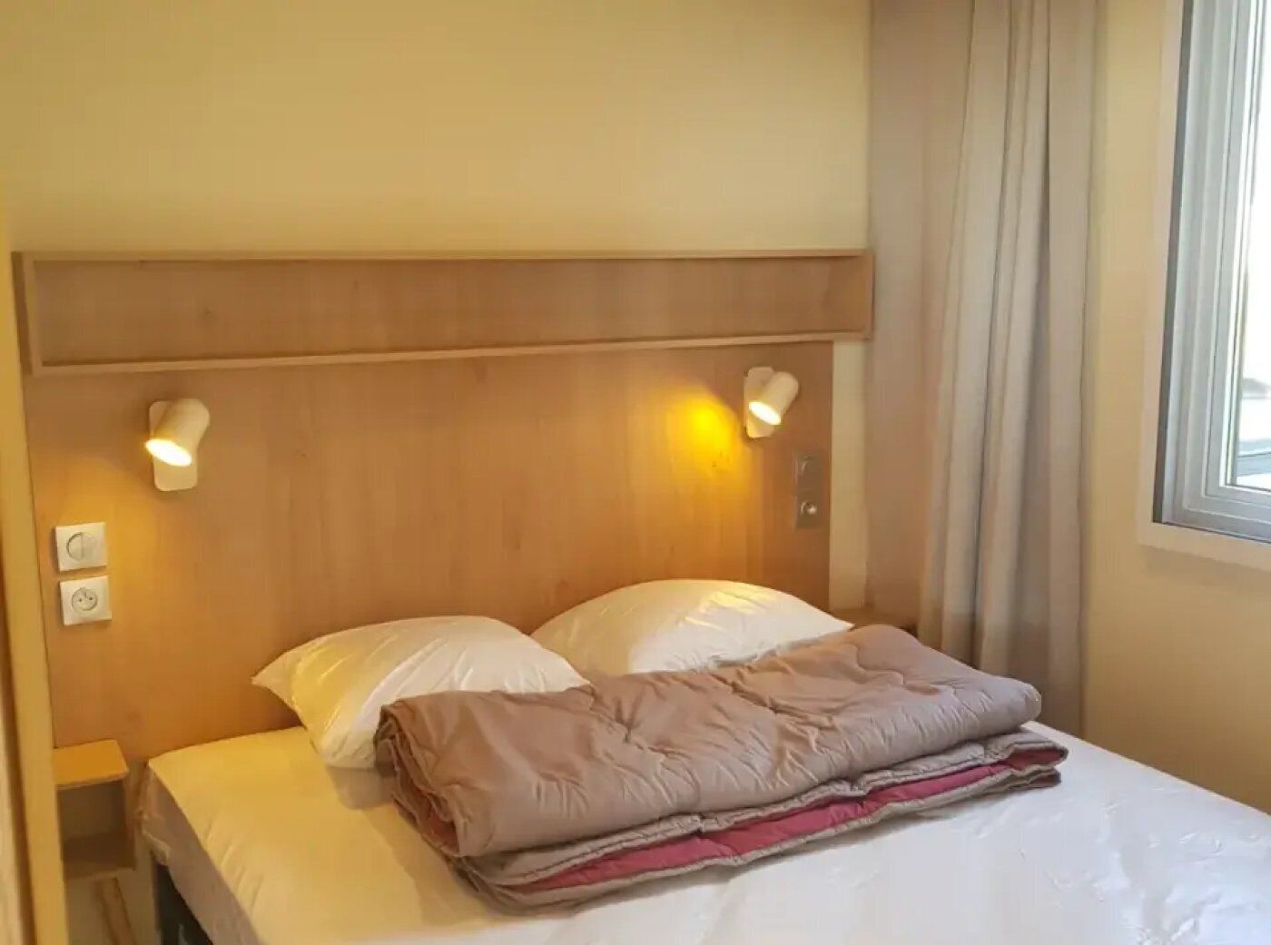 A hotel room with a wooden headboard, two bedside lamps, a bed with white sheets and a folded blanket, and a window with curtains.