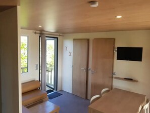 A room with a sliding glass door, a flat-screen TV mounted on the wall, and a wooden table with chairs. - Camping 3 étoiles - Piscine  - ccbffde (Arces)