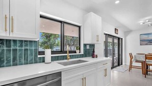 A modern kitchen with white cabinets, a stainless steel dishwasher, a window with a view, and a dining area with a table and chairs. - Casa Mimosa Family Tropical Oasis with Pool (Miami)