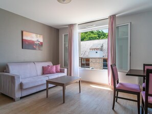 A room with a sofa, a table, and a chair, a painting on the wall, and a view of a house through a window. - Art Village Apartment for Four (Pont-Aven)