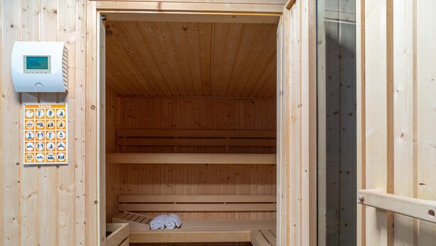 Sala de sauna com bancos de madeira, termostato digital e placa indicativa de sauna.