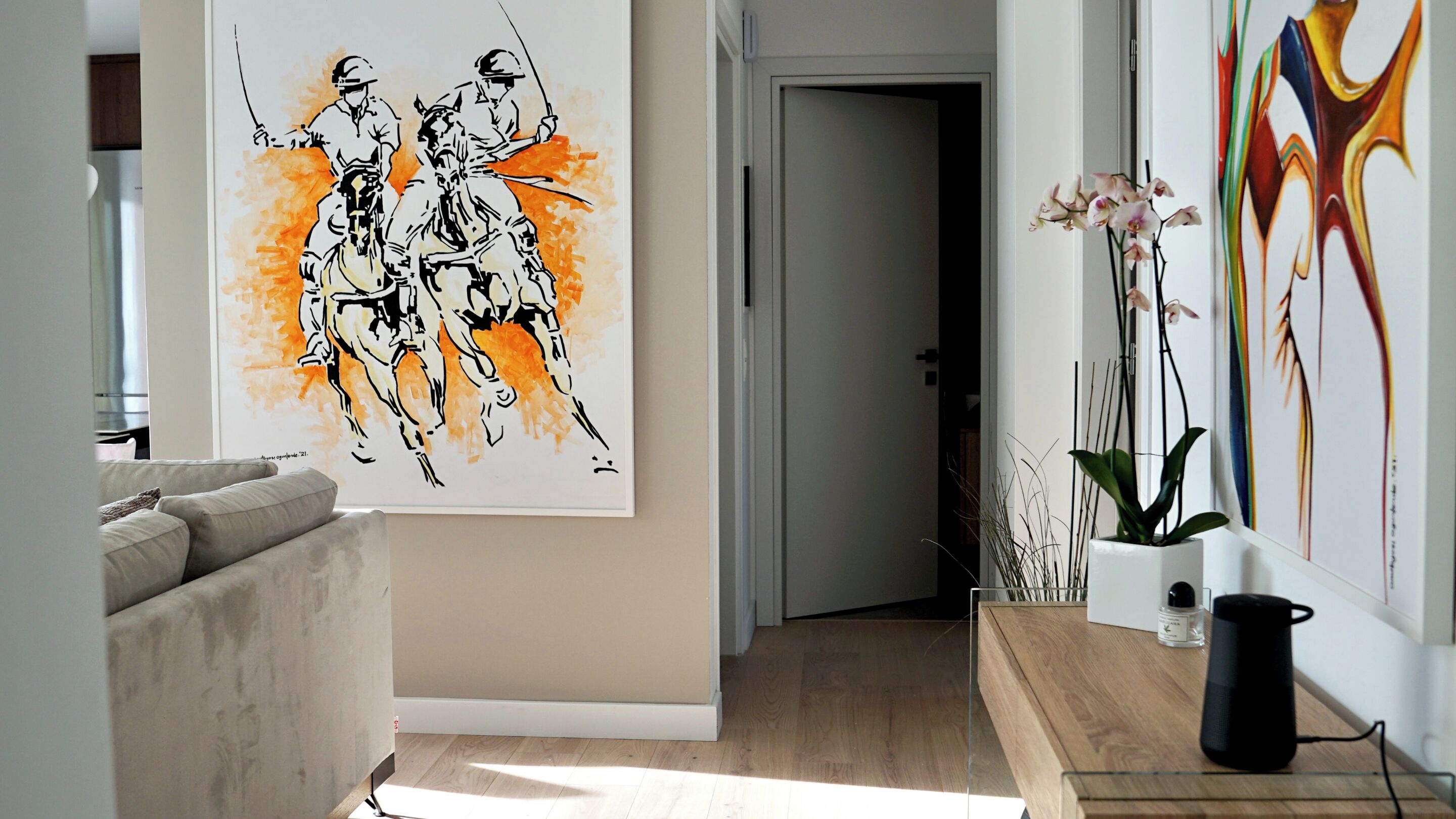 A hallway with a painting of horse riders, a sofa, a potted plant, and a coffee maker on a table.