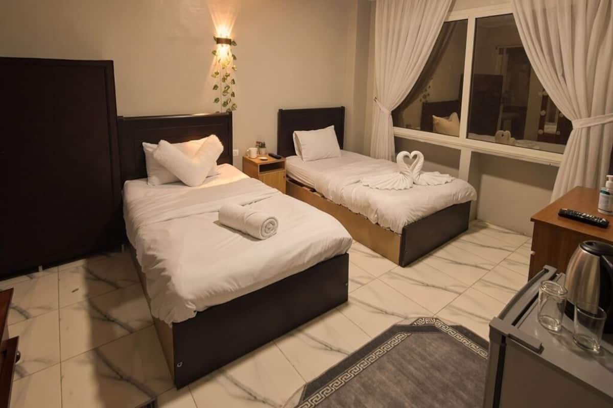 Una habitación de hotel con dos camas, una mesita de noche, un escritorio con hervidor y una ventana con cortinas, además de una lámpara de pared.