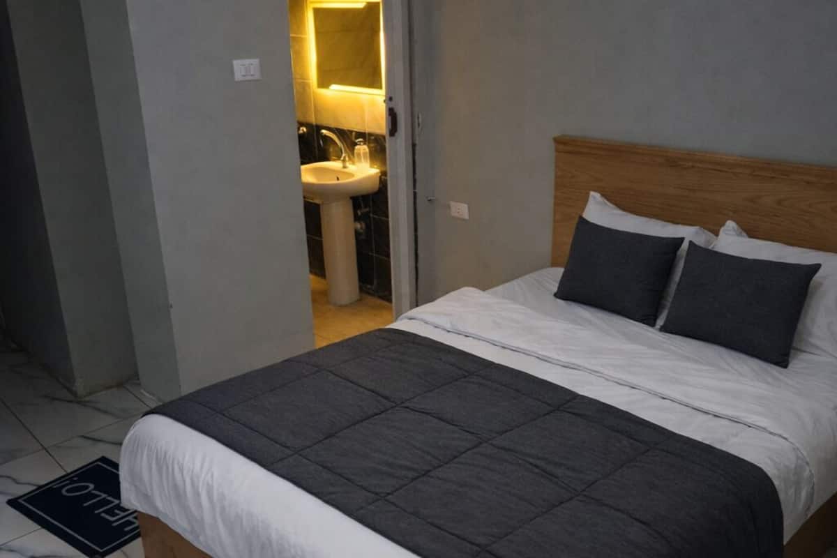 Un dormitorio con una cama, cabecera de madera, mesita de noche y un baño visible a través de una puerta abierta.