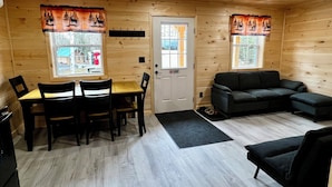 Een houten chalet van binnen met een eettafel, stoelen, een bank en twee ramen met gordijnen.