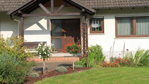 Una casa con puerta marrón, un pequeño porche y un jardín lleno de plantas y flores.