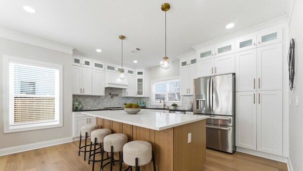 Cocina moderna con armarios blancos, una isla central con taburetes, electrodomésticos de acero inoxidable e iluminación colgante.