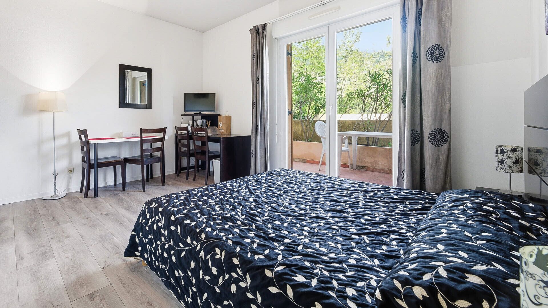 Une chambre à coucher avec un lit, un bureau, des chaises, une télévision et une vue sur la verdure par la fenêtre.