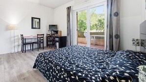 A bedroom with a bed, a desk, chairs, a TV, and a view of greenery through the window. - Charming Studio - Sleeps 2 - bright (Grasse)