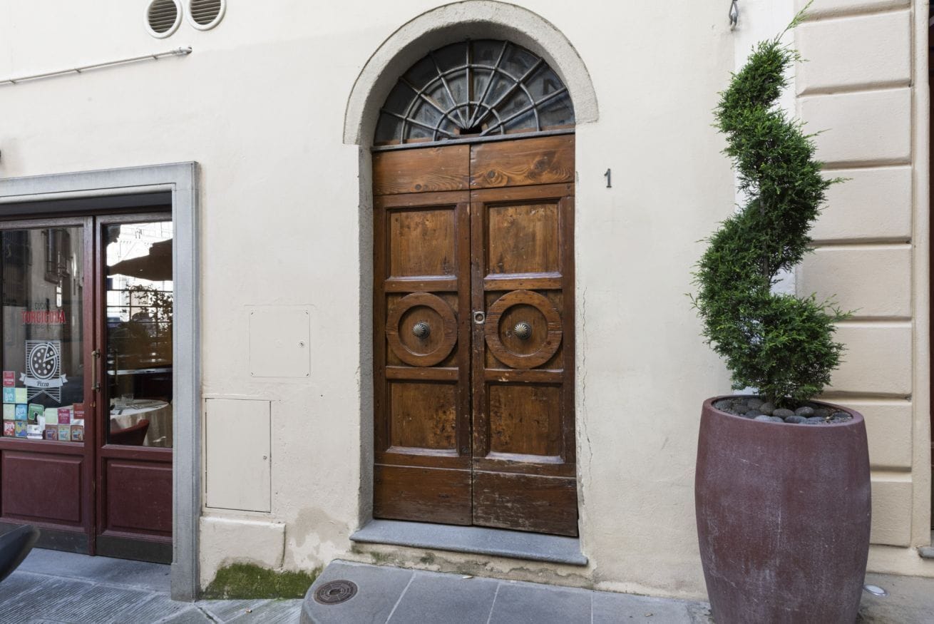 A wooden double door with a decorative arch above it, a potted plant beside the door, and a building number '1' on the wall.