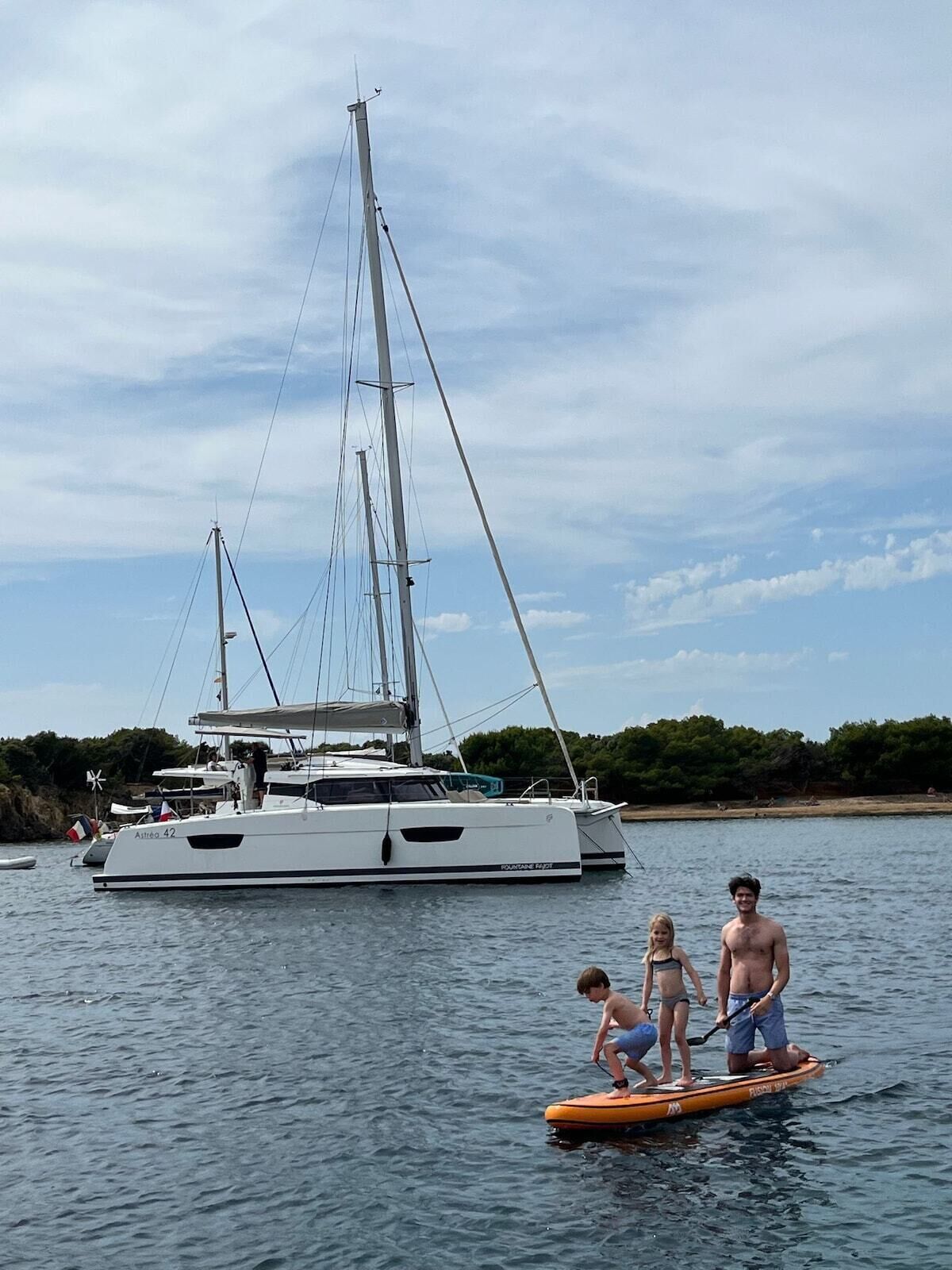 Un velero con camarote y una persona de pie en una tabla de remo en el agua.