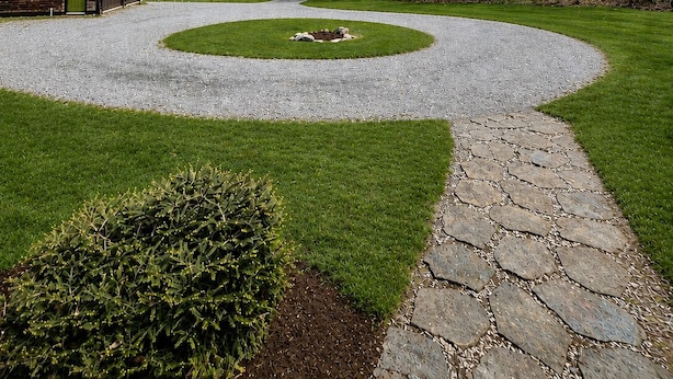 Un camino de acceso circular que conduce a una construcción de madera con una puerta verde, rodeada de un césped cuidado y árboles.