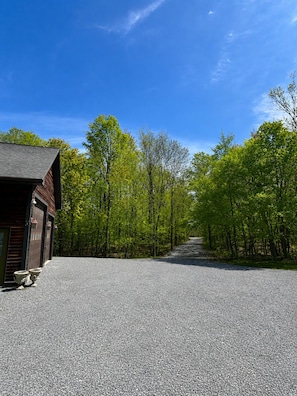 Une allée de gravier mène à un bâtiment en bois doté d’un garage, le tout étant entouré d’arbres.