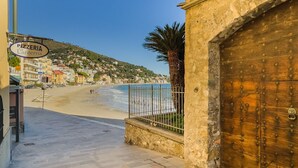 A coastal street with a wooden door, a sign for a pizzeria, and a beach in the background.