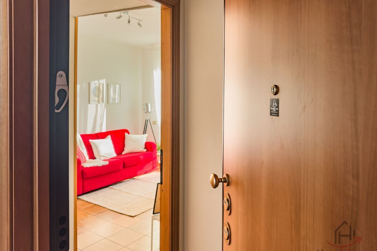 Habitación de hotel con puerta de madera, sofá rojo y pared blanca con un letrero pequeño.