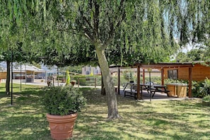 Un pavillon en bois avec des tables de pique-nique, un grand arbre et une plante en pot, le tout situé dans un espace verdoyant.