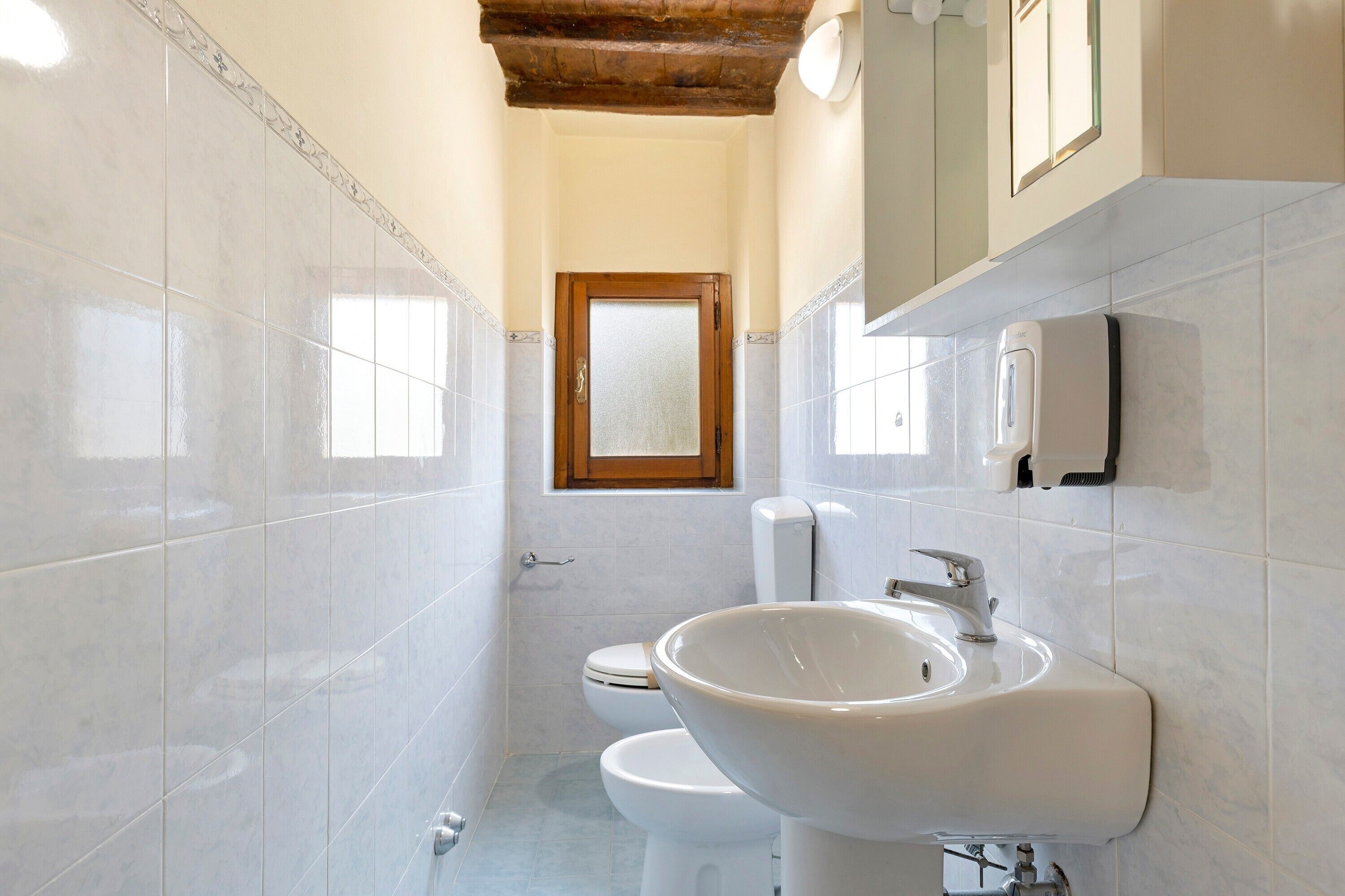 A bathroom with white tiles, a wooden door, a toilet, a sink, and a soap dispenser.