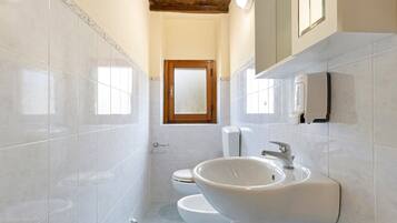 A bathroom with white tiles, a wooden door, a toilet, a sink, and a soap dispenser.