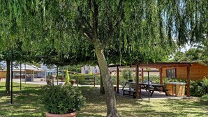 A wooden pavilion with picnic tables, a large tree, and a potted plant in a grassy area.