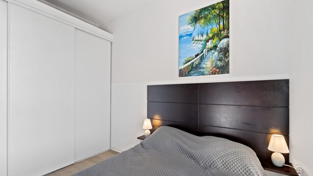 A bedroom with a bed, a headboard, a nightstand with a lamp, a painting on the wall, and a wardrobe. - Apartment Near the Center of Grasse (Grasse)