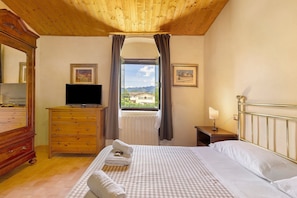 Une chambre à coucher avec un lit, une commode, un téléviseur, une fenêtre avec des rideaux et deux tableaux encadrés accrochés au mur.