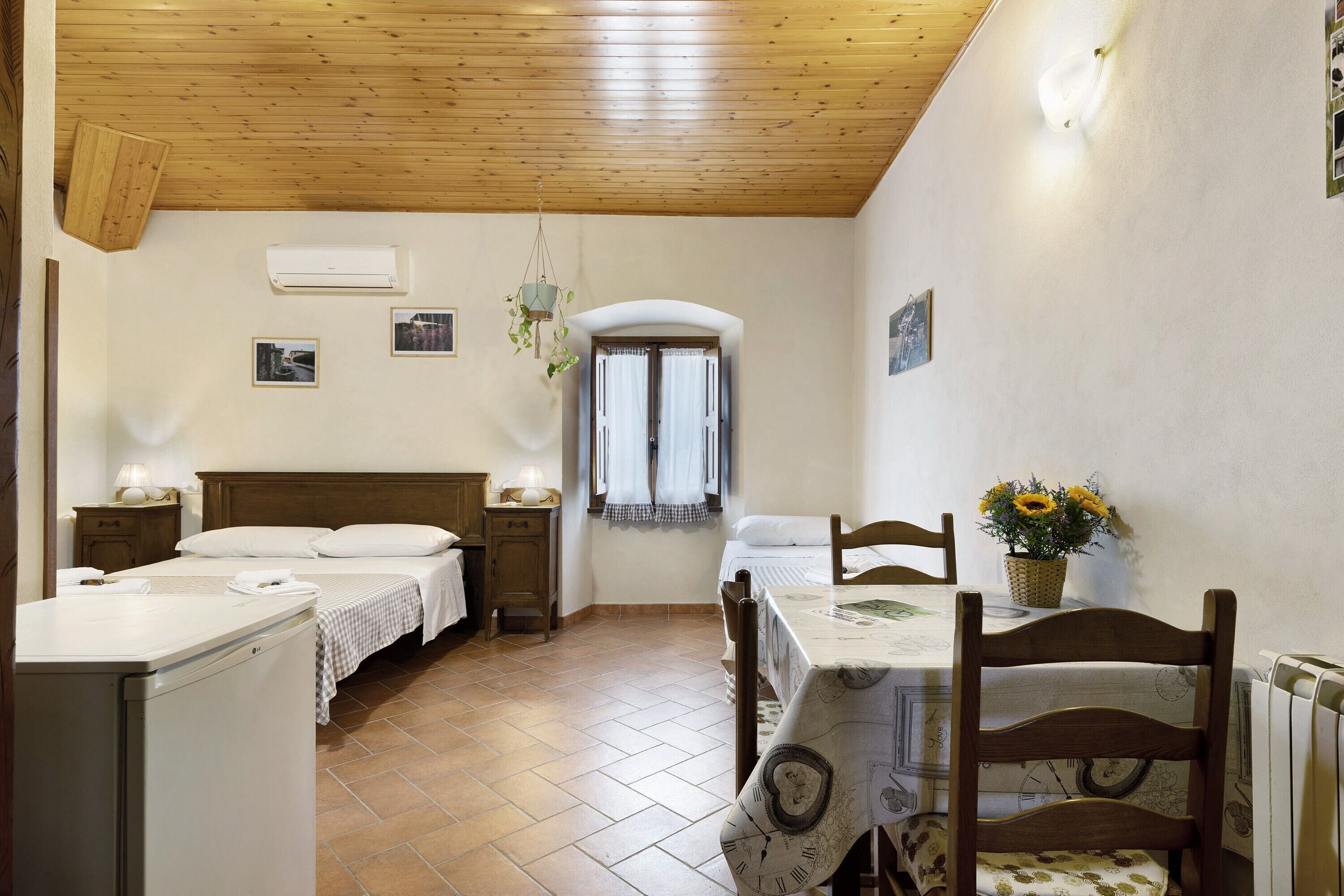 A bedroom with a wooden ceiling, a bed with white linens, a bedside table with a lamp, and a window with curtains.