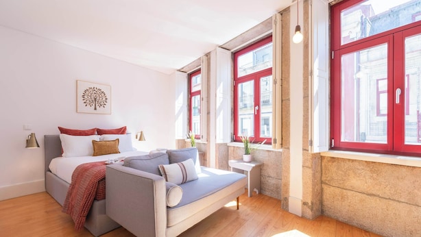 Ein modernes Schlafzimmer mit einem Bett, einem Schlafsofa, einem kleinen Tisch und einem Fenster mit roten Lamellen.