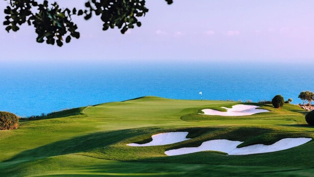 A golf course with bunkers and a flagstick, overlooking a body of water.