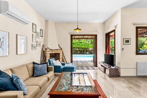 A living room with a beige sofa, blue armchairs, a glass coffee table, a fireplace, a TV, and a view of a swimming pool.