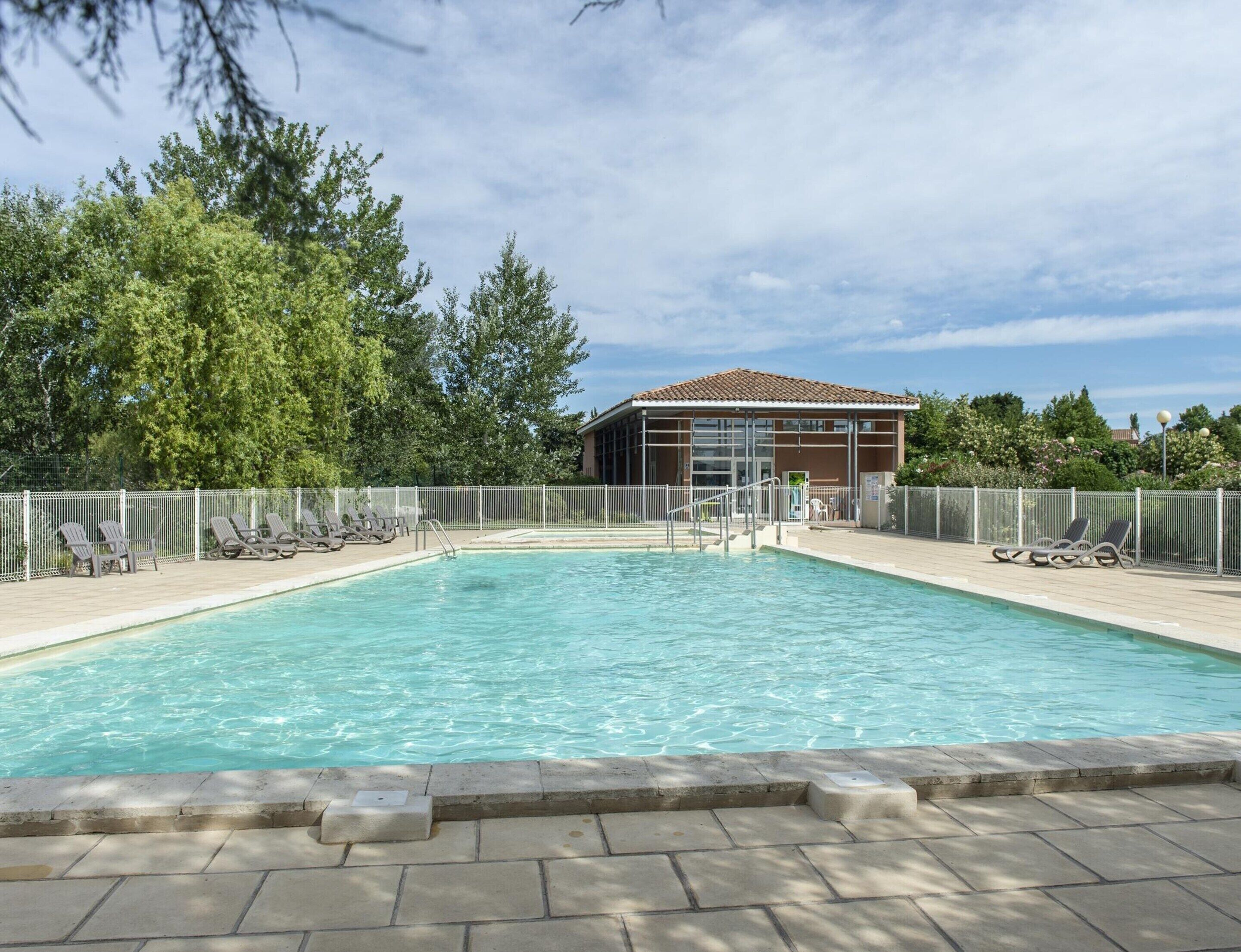A swimming pool with a tiled deck, surrounded by a fence, and a building with a tiled roof in the background.