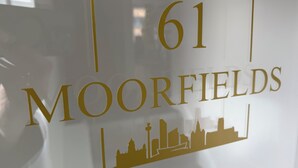 Segnale con il numero 61 e il nome 'MOORFIELDS' in caratteri dorati, con la sagoma di edifici sottostante.