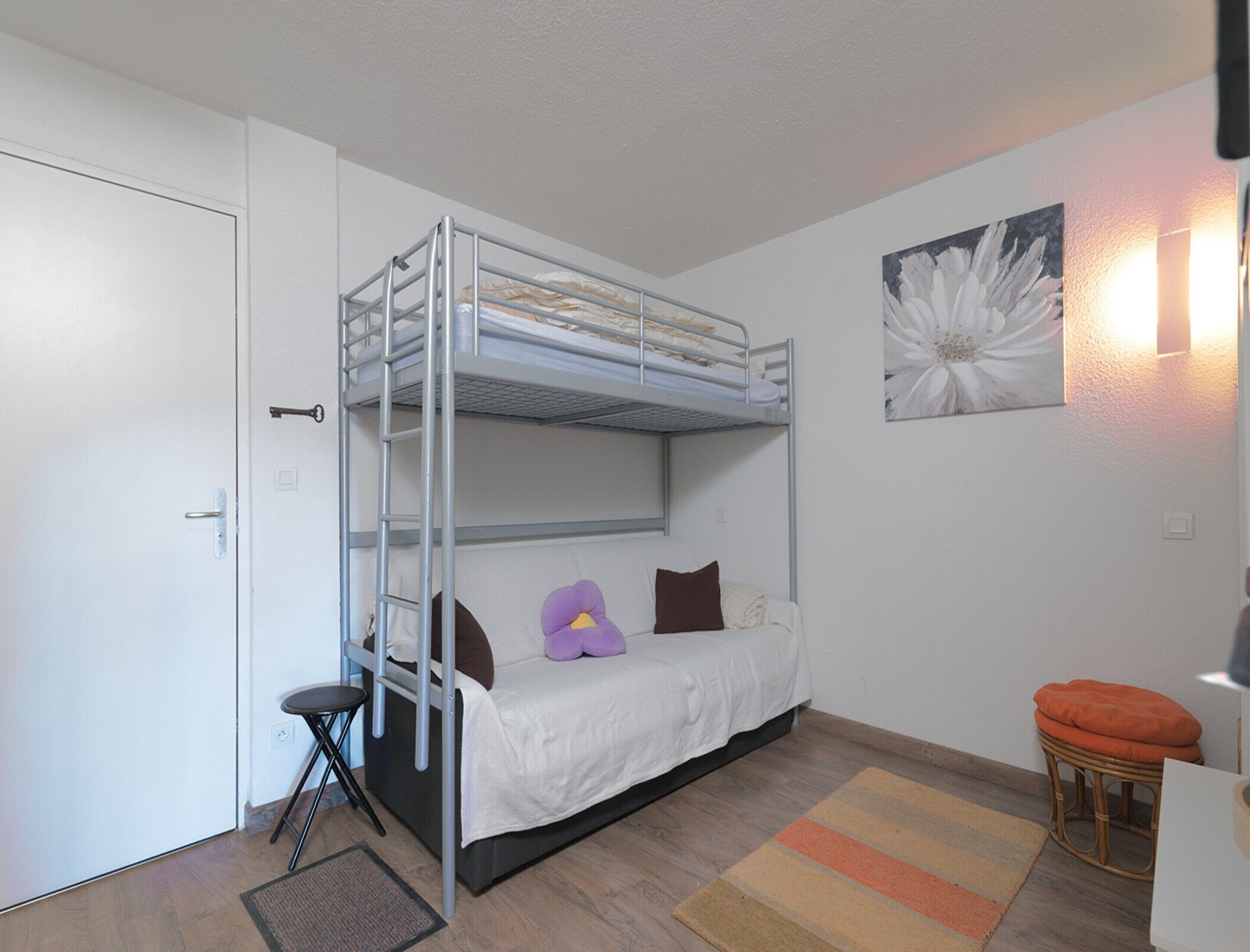 A bunk bed room with a door, a small stool, a rattan chair, and a wall-mounted light fixture.