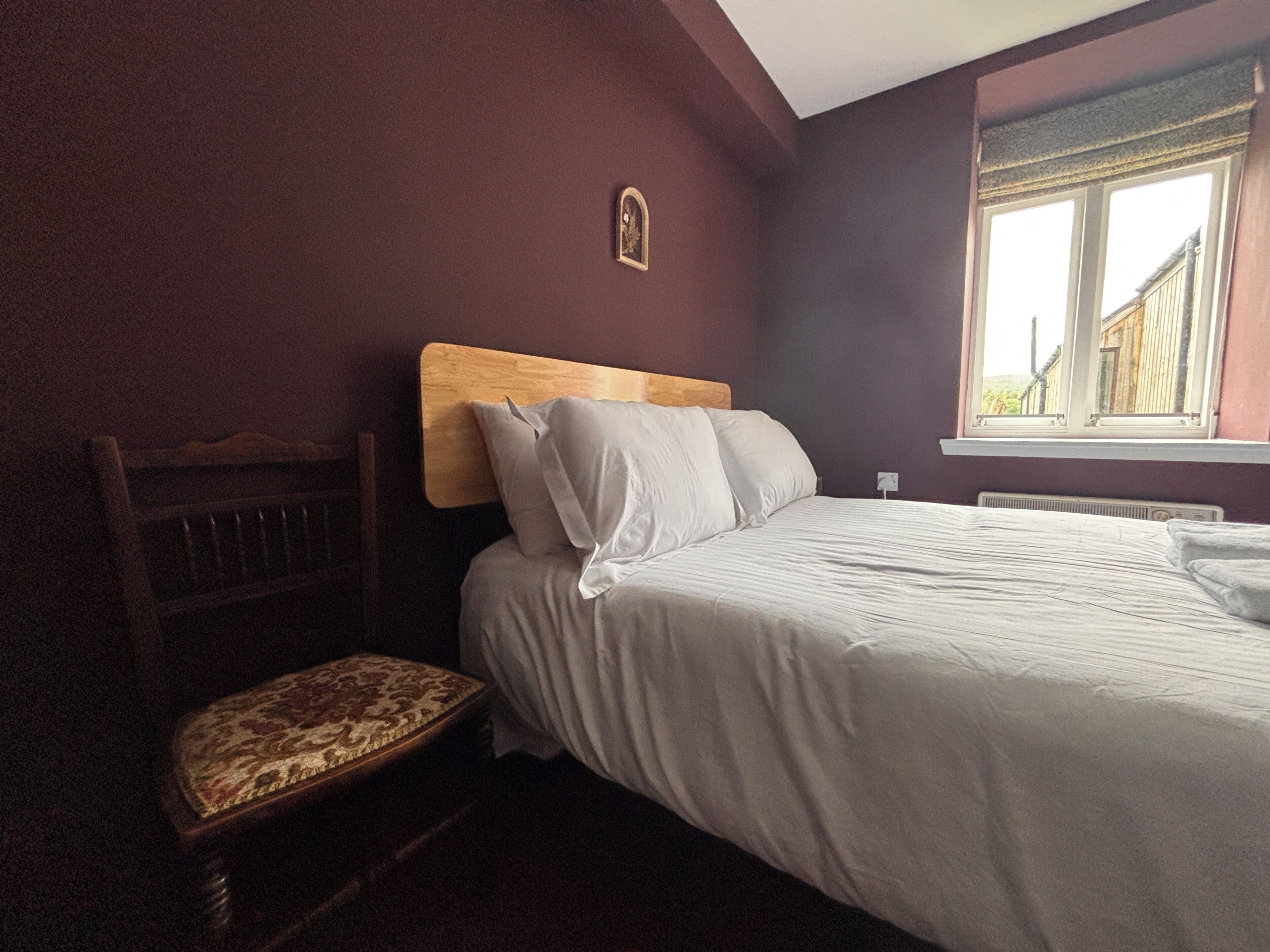 A bedroom with a wooden headboard, a single bed, a bench with a patterned seat, and a window with a view of buildings.