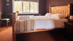 A bedroom with a wooden bed, a window with a view of trees, a chair, and a bedside table with a lamp.