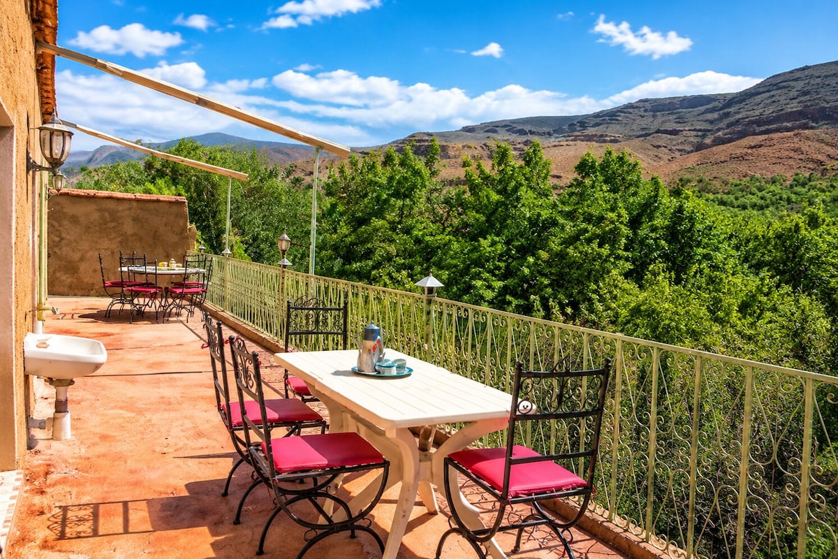 An outdoor terrace with a table and chairs, a view of green hills, and a clear sky.
