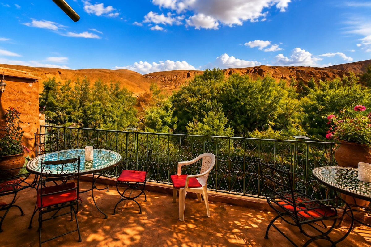 A terrace with two round tables and chairs, surrounded by a metal railing, overlooking a mountainous landscape with trees and a clear sky.