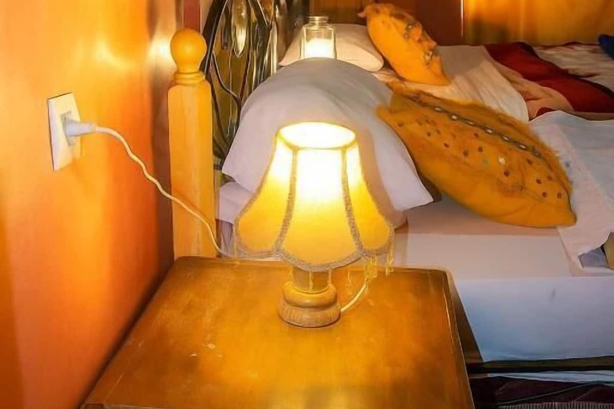 Una habitación con una cama, una lámpara de noche, una mesita de noche de madera y un ventanal con cortinas.