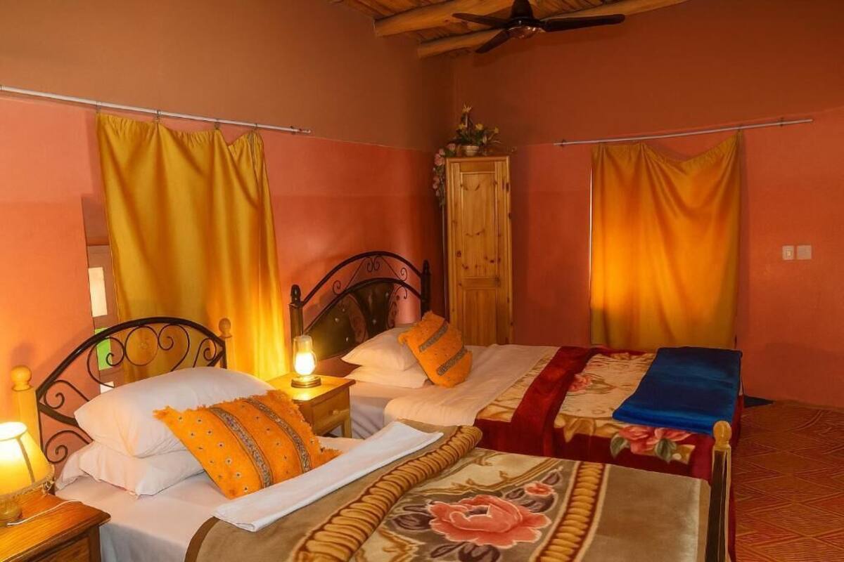 Una cama doble con una colcha estampada, una mesita de noche con lámpara y un armario de madera en una habitación con paredes naranja y cortinas amarillas.