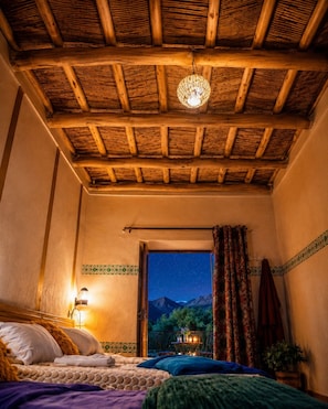 Una habitación con techo de madera, una cama con almohadas, una mesita de noche con lámpara y vistas a montañas a través de una ventana.