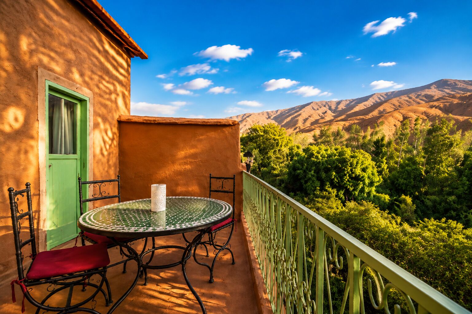A balcony with a round table and chairs, a green door, and a view of mountains and greenery.