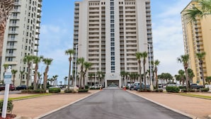 Un grand hôtel avec une entrée centrale, entouré de palmiers et de voitures garées.