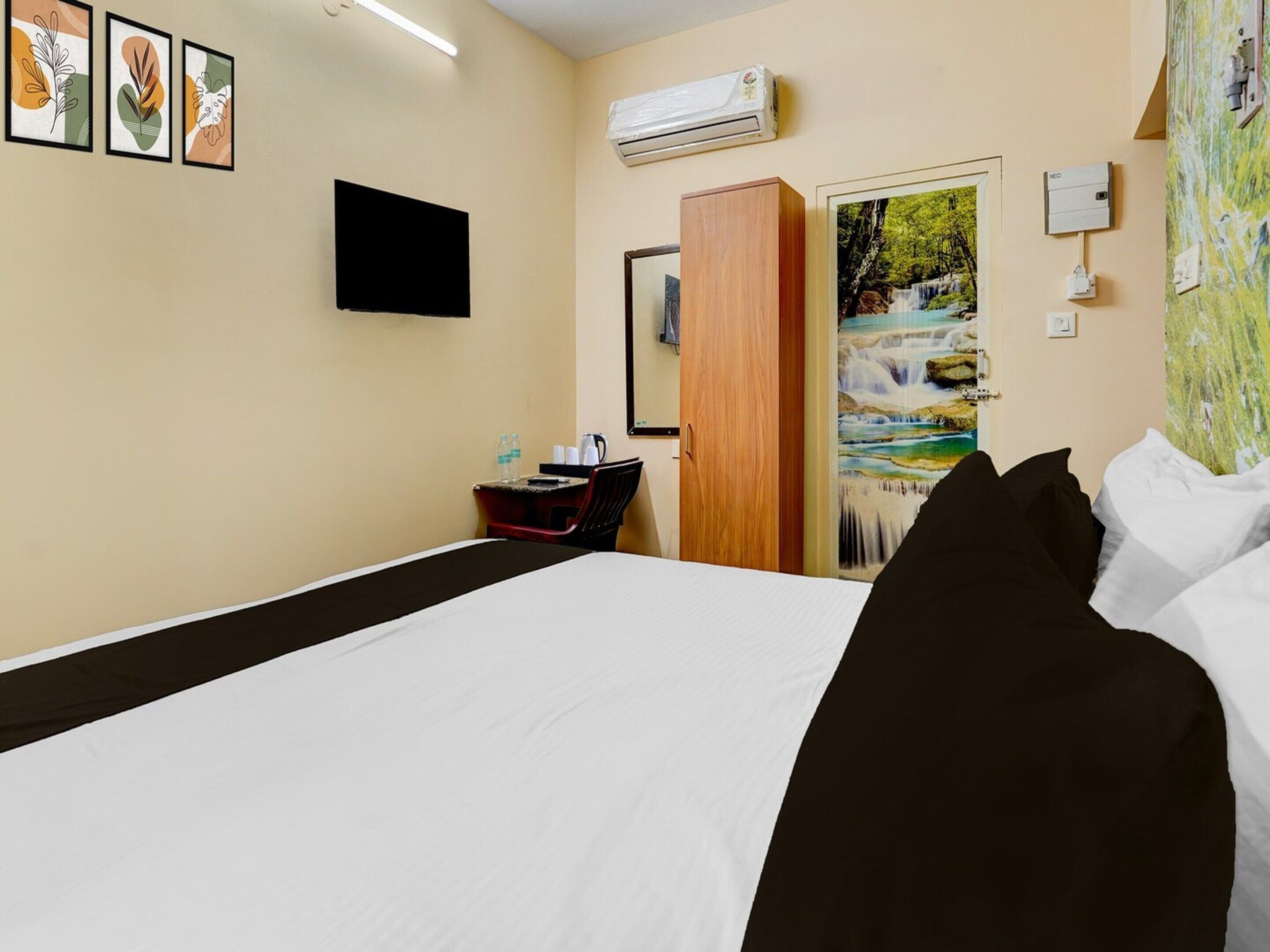 A hotel room with a bed, a desk with a chair, an air conditioner, a painting of a waterfall, and a television on the wall.