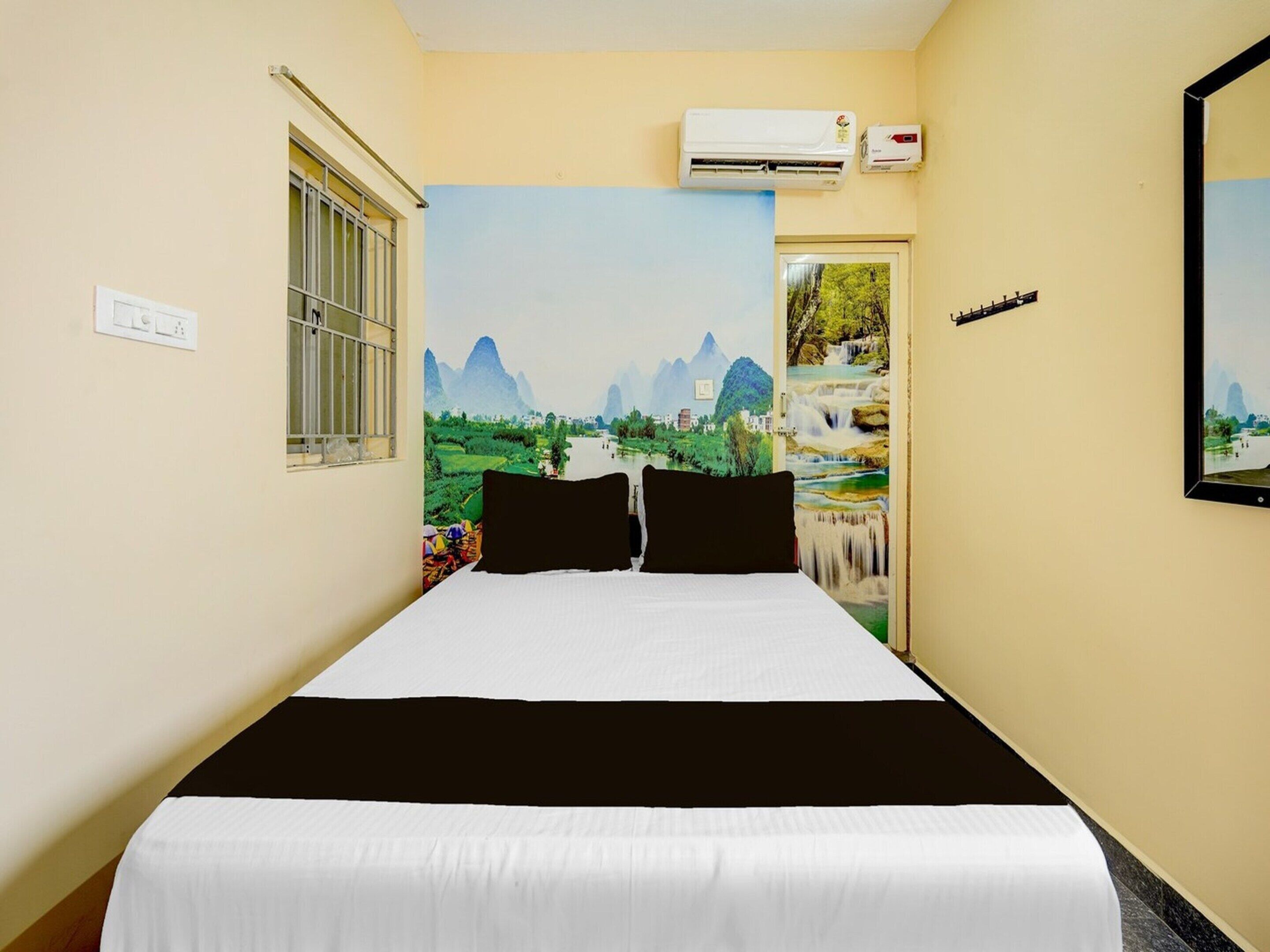 A hotel room with a bed, an air conditioning unit, a window with bars, and a mural of a landscape.