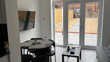 A modern living room with a dining area, large sliding glass doors to a backyard, and a flat-screen TV mounted on the wall.