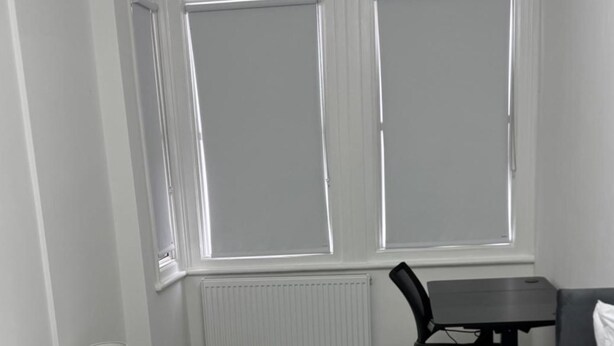Quarto com uma janela grande, um radiador, uma cadeira preta e uma mesa pequena.