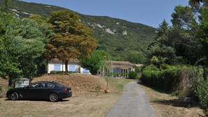 Un camino rural con un coche estacionado, una casa con techo de tejas y una colina al fondo.