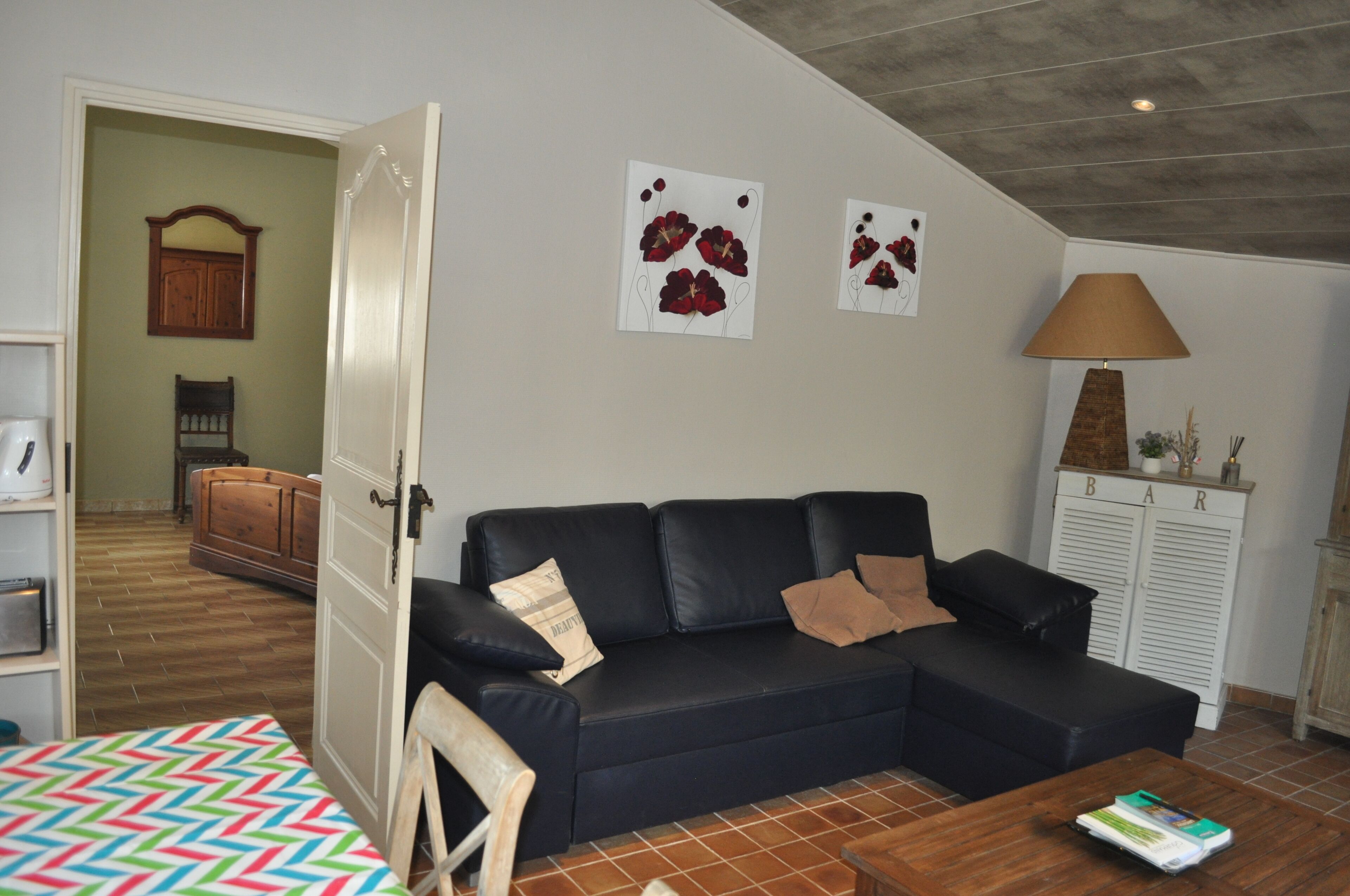 Een woonkamer met een zwarte bank, een houten salontafel, een lamp en een deur die naar een andere kamer leidt.