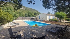Una piscina con sillones de descanso, un muelle de madera y una casa de fachada blanca con contraventanas azules.